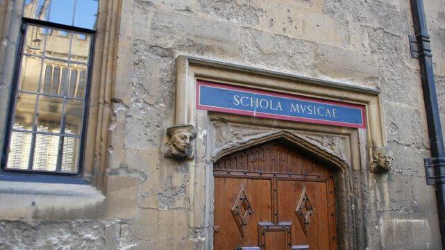 Doorway detail at Oxford University showing Schola Musicae meaning school of music, with sculpted stone faces, aged wall textures, and leaded glass window in close view.