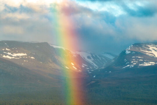 Aerial view of a vibrant rainbow appearing over the snow-capped peaks and rugged valleys of Cairngorms National Park Scotland, United Kingdom.