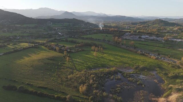 Cinematic aerial shot of lush green fields, mountains, and a small pond in the Italian landscape during a warm sunset. Great for travel and nature documentaries.