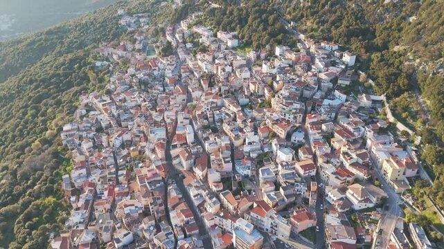 Cinematic aerial shot of the historic town of Baunei nestled in the Supramonte mountains of Sardinia, Italy, showcasing authentic Mediterranean architecture at sunset.