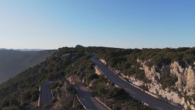 Cinematic aerial shot of a serpentine road through the rugged mountains of Baunei, Sardinia, during golden hour. Perfect for travel, adventure, and automotive content.
