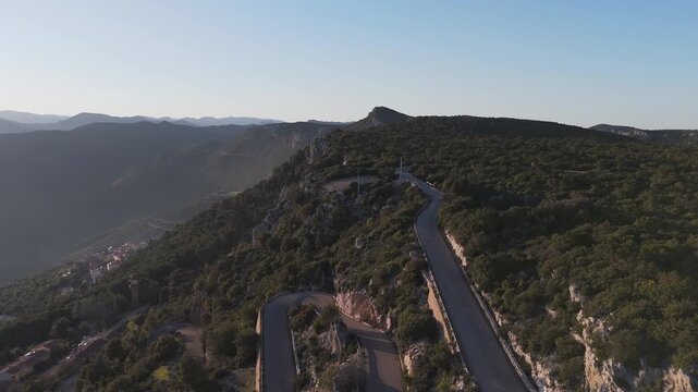 Cinematic aerial shot of a serpentine road winding through the rugged mountains of Baunei, Sardinia, captured during the warm golden hour light.