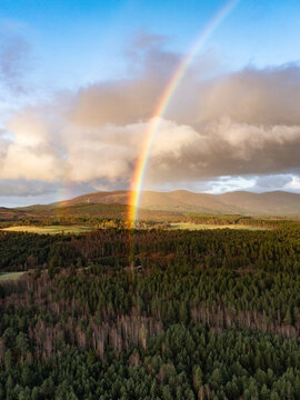Aerial view of Cairngorms National Park with a vibrant rainbow over dense evergreen forests and rolling mountain peaks under a cloudy sky Scotland, United Kingdom.