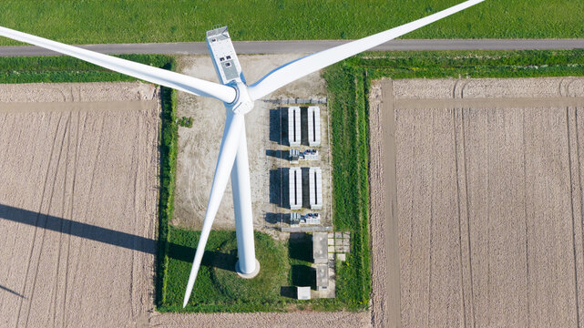 Aerial view of a large white wind turbine and battery storage units on a green plot surrounded by plowed agricultural fields in Ooltgensplaat, Zuid-Holland, Netherlands.