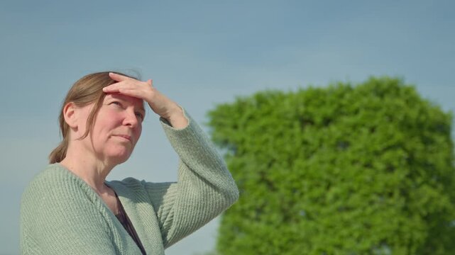Middle-aged woman outdoors shields her eyes and face from bright sunlight as she looks into the distance beside a neatly trimmed green hedge, 4K