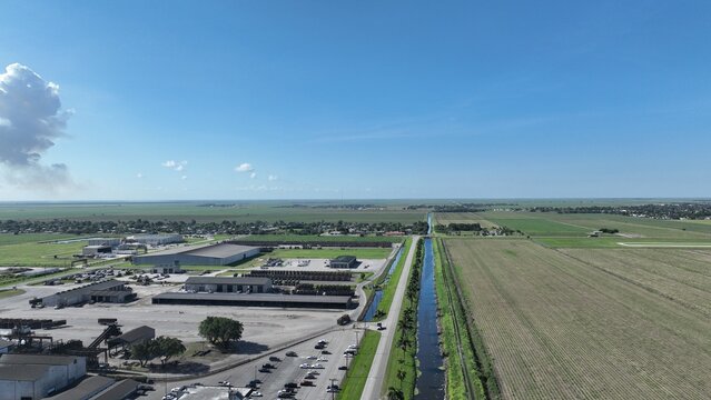 Aerial view of large industrial warehouses and vast agricultural fields divided by a straight water canal under a blue sky with a large white cloud.