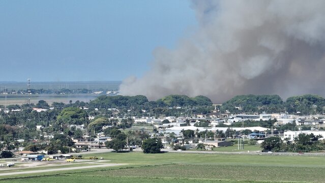 Aerial view of a massive plume of dark smoke rising from a fire behind a suburban area with green trees and industrial buildings.