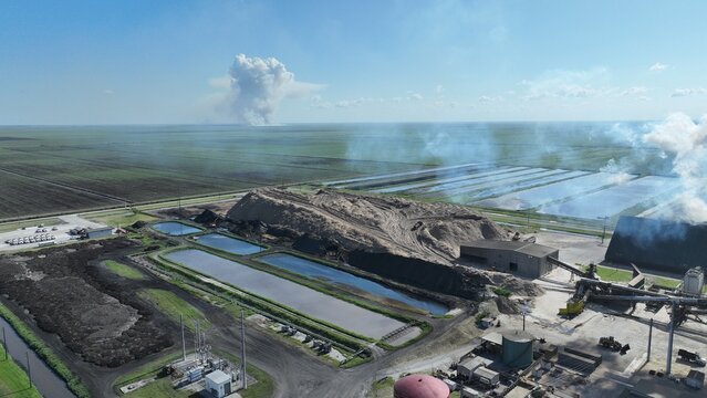 Aerial view of an industrial processing plant featuring settling ponds and large material piles with smoke plumes rising from the facility and distant agricultural fields.