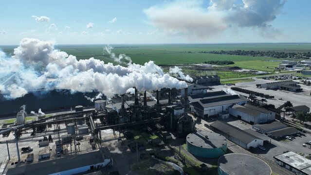 Aerial view of a large industrial sugar mill facility with thick white smoke rising from chimneys into a blue sky over green agricultural fields.