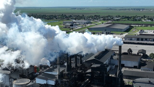 Aerial view of an industrial factory with thick white smoke billowing from tall chimneys over green fields, warehouses, and a rural landscape.
