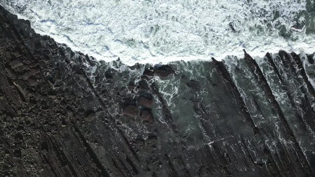 Aerial top down view of ocean waves breaking on the unique flysch rock formations of Sakoneta beach in the Basque Country, a mesmerizing display of nature's geological power and beauty