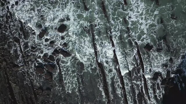 Dramatic aerial topdown view showing powerful waves crashing against the unique Flysch rock formations of Sakoneta beach in the Basque Country, creating a mesmerizing natural spectacle