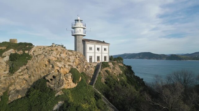 Aerial view of the historic Getaria lighthouse perched on a rocky cliff, with the vast ocean in the background and a staircase leading up to the historic building on a sunny day