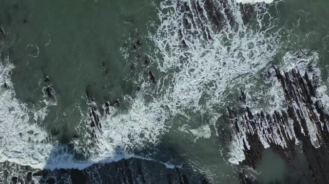 Aerial top down perspective of powerful ocean waves crashing against the unique Sakoneta flysch rock formations along the Basque Coast Geopark, creating mesmerizing white foam patterns