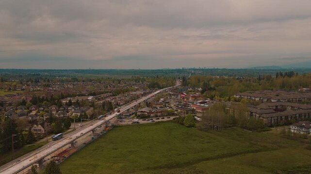 Expansive high altitude view showing busy suburban highway traffic flowing near active elevated concrete transit guideway construction site bordered by residential neighborhoods under cloudy sky orbit