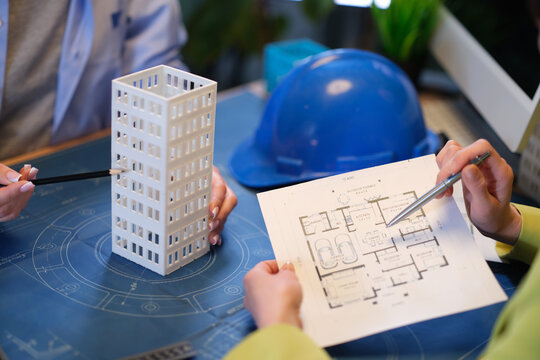 Architects reviewing building model and floor plan, hands pointing at blueprint with pen, scale model of multi story building,