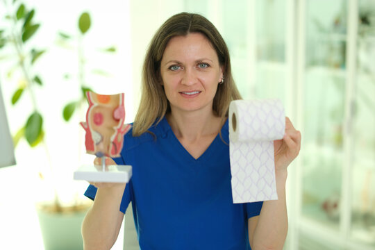 Caucasian female physiotherapist holding rectum model and tissue roll in bright clinic setting, wearing blue scrubs, smiling,