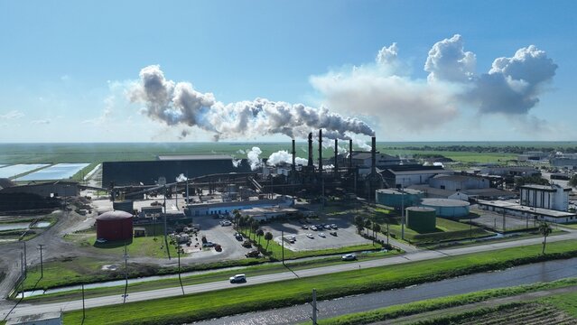 Aerial view of a large industrial factory complex with smokestacks emitting white plumes under a clear blue sky.