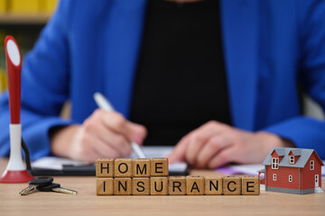 Obraz premium A professional is seated at a desk focused on taking notes about home insurance. On the table, wooden blocks spell out home insurance next to a small model house and key set.
