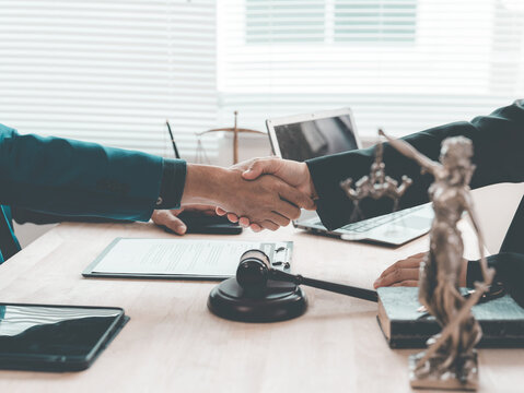 The lawyer is shaking hands with his client to congratulate them on the successful settlement of a legal case, with a gavel and legal scales beside them.