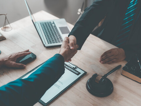The lawyer is shaking hands with his client to congratulate them on the successful settlement of a legal case, with a gavel and legal scales beside them.