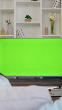 Rear View. Family couple watches green screen TV mockup sitting on couch in living room together. Man and woman watching TV chroma key in domestic cinema looking TV show or news in home. Slow motion.