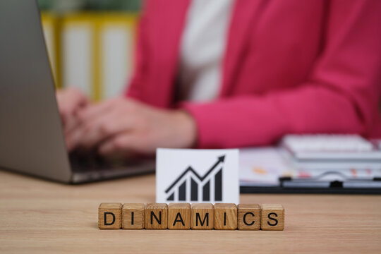 A professional woman works at a laptop in a contemporary office, analyzing growth charts. Wooden blocks spelling Dinamics sit in front, symbolizing strategic planning and progress.
