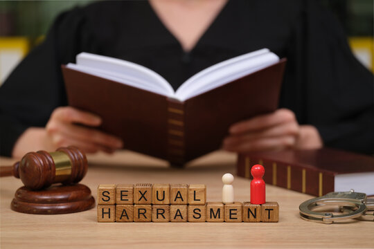 In a solemn courtroom, a judge reads legal texts about sexual harassment. Wooden blocks spell the term, while a gavel and figurines symbolize justice and advocacy for victims.