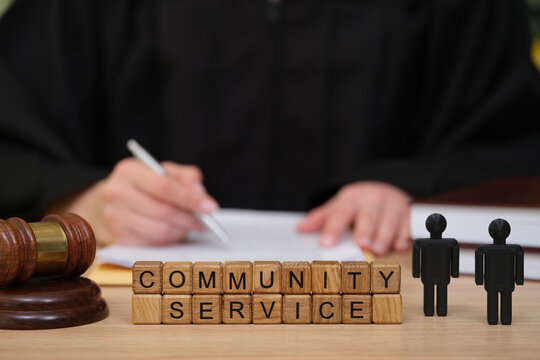 In a courtroom setting, a judge in black robes attends to paperwork related to community service. Wooden blocks spell out community service next to a gavel and figurines.