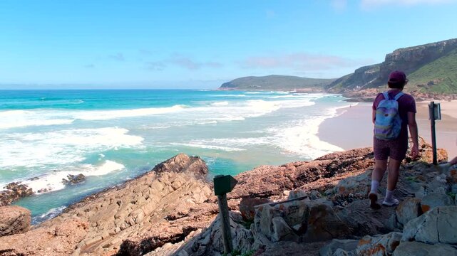 Hikers on Robberg hiking trail walking over jagged rocks with clear ocean waves to side running out onto beach in Plettenberg Bay