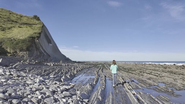 Young adventurous woman walking toward the ocean on the impressive Flysch Route, a unique geological formation on the coast of the Basque Country between Zumaia and Deba on a sunny day