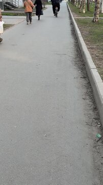 Rear view sidewalk with cyclist and passers walking on spring day along narrow paved footpath by curb and trees, shoppers