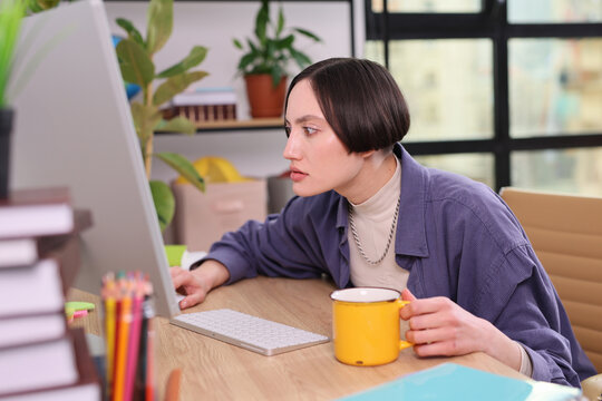 With a hint of curiosity, a woman studies her computer screen intently while cradling a yellow mug in a stylish, bright workspace filled with plants and books.