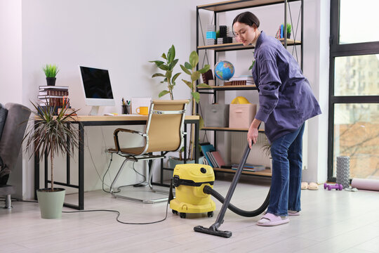 In a bright and modern office, a woman enthusiastically cleans the floor with a yellow vacuum cleaner, creating a tidy and inviting workspace full of greenery.