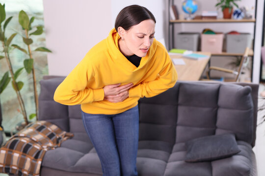 A young woman sits on a sofa, cradling her stomach in discomfort. Sunlight pours into the room as she experiences a moment of pain, surrounded by a warm atmosphere.