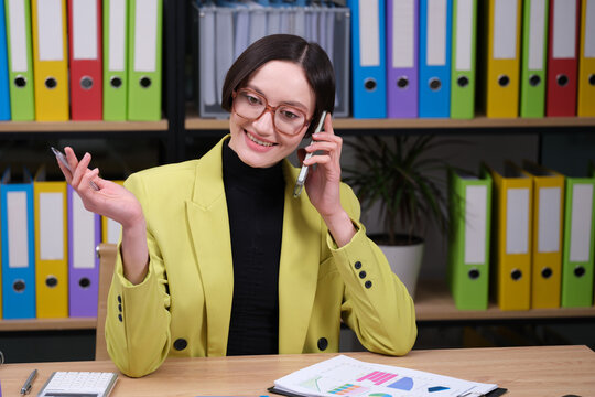A woman dressed in a bright yellow blazer engages in a phone conversation. Colorful binder folders fill the background, adding energy to the workspace she is in.