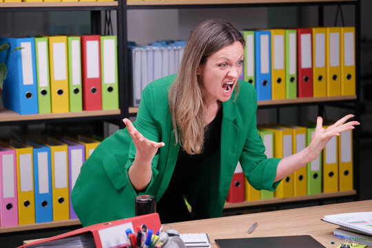 In a vibrant office filled with colorful folders, a woman in a green blazer expresses intense frustration. Her animated gestures capture the chaos of a demanding work environment.