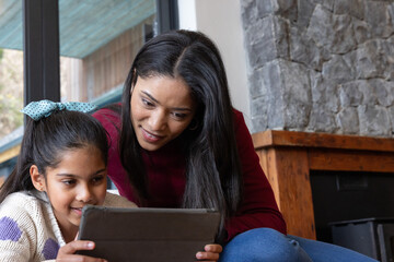 Naklejka premium Asian mother and daughter sitting on couch in living room, leaning, holding tablet with folio case