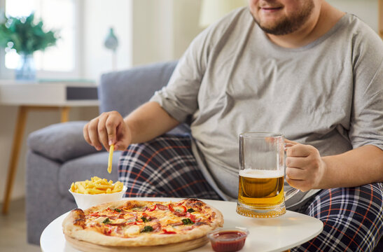 Overweight man eating fries, pizza and drinking beer at home. Plus size funny guy sitting on living room sofa, holding mug and potato chip, making choice of unhealthy habit, fast food diet and obesity