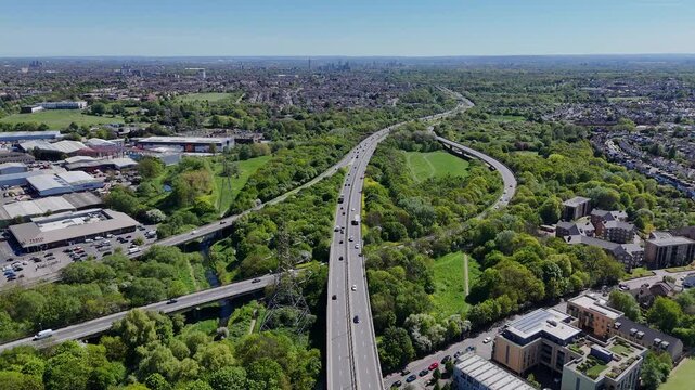 Aerial drone view of a major highway interchange with moving traffic surrounded by residential areas and green spaces on a clear day.