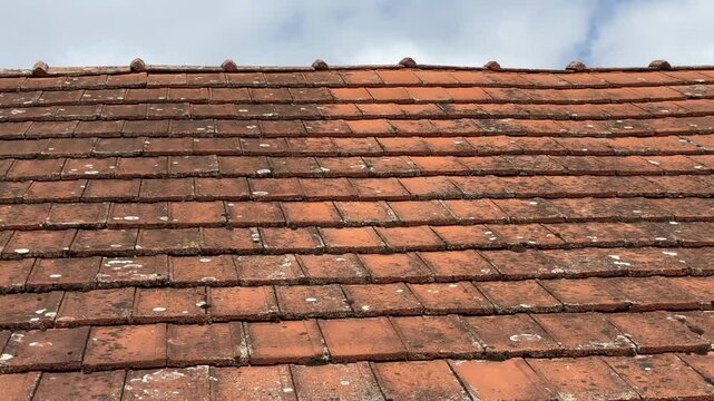 Close-up tracking shot of weathered red clay roof tiles on a small house, camera gliding over textured rooftop under cloudy sky, showcasing rustic architecture and aged surface detail