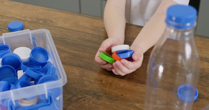 Young boy holds colorful bottle caps near clear container and plastic bottle on table. Child sorts small lids with hand preparing pieces for recycling box slow motion