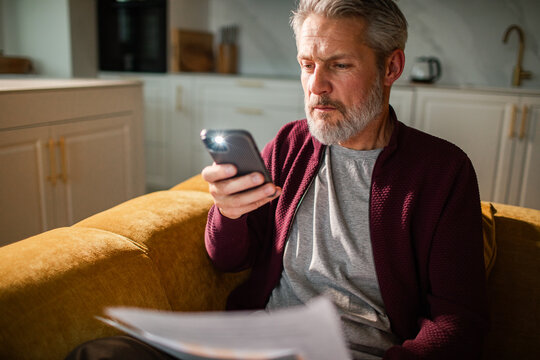 Older man using smartphone flashlight to read bills at home