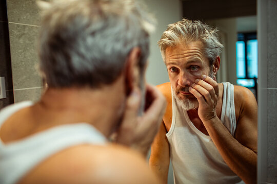 Middle-aged man examining eye bags in bathroom mirror at home