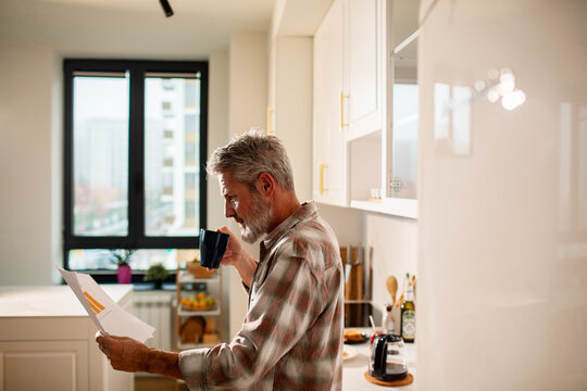 Older man reviewing bills while drinking coffee in home kitchen