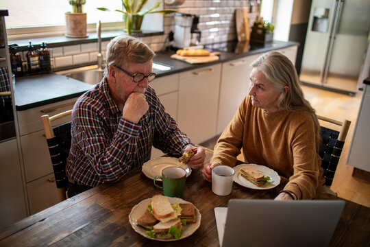 Senior couple eating lunch and using laptop in home kitchen