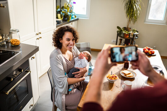 Smiling mother holding newborn while being photographed in home kitchen