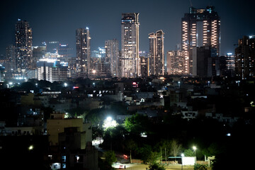Fototapeta premium Modern highrise multi storey skyscrapers at night with lights shining through with smaller independent houses in foreground showing the income disparity in cities like gurgaon, delhi, bangalore, hyder