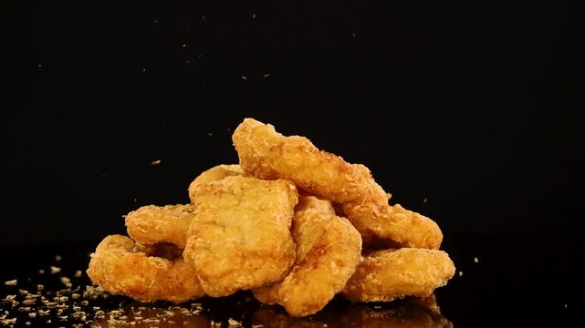 Slow motion rotation of crispy chicken nuggets with falling spices on black background.
