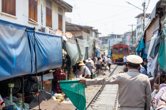 Focused railway official holding green flag directing approaching train through bustling crowded outdoor market on narrow track managing safety of local vendor and tourist